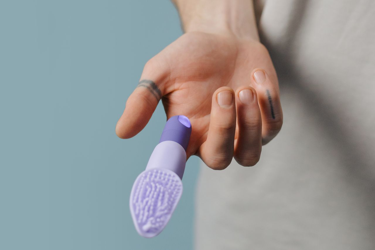 gallery-5 A close-up image showing a hand holding a purple silicone finger vibrator against a neutral background.